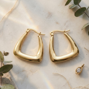 Geometric gold hoop earrings by Manadh Jewels crafted from premium stainless steel with a polished gold finish, displayed on a marble pedestal alongside a model wearing the earrings in a luxury setting