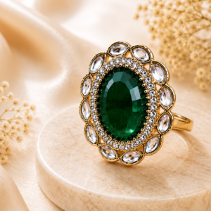 Vintage emerald green statement ring with an oval gemstone surrounded by sparkling crystal accents in a gold-tone finish, displayed on a black marble pedestal by Manadh Jewels.