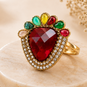 Vintage ruby-red statement ring with a triangular center stone, multicolor gemstone accents, crystal halo, and gold-tone finish displayed on a black marble pedestal by Manadh Jewels.