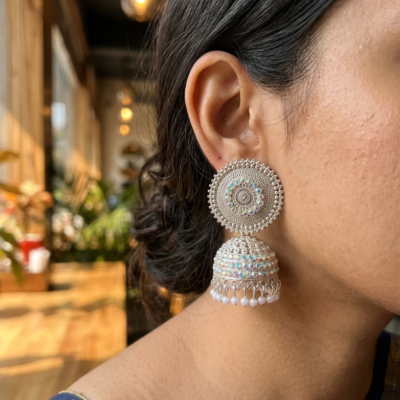 Premium oxidized jhumka earrings with intricate circular detailing, shimmering stones, and pearl drops displayed on a beige marble surface by Manadh Jewels