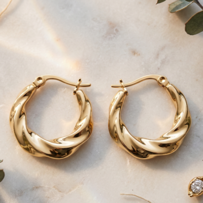 Twisted gold hoop earrings by Manadh Jewels crafted from premium stainless steel with a polished gold finish, displayed on a marble platform in an elegant luxury setting
