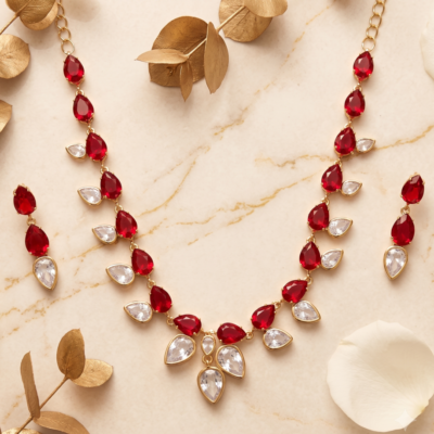 Elegant red gemstone necklace and matching earrings with teardrop-cut red and clear stones set in gold-toned metal, arranged on a luxurious marble background with golden leaf accents.