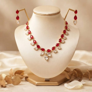 Elegant red gemstone necklace and matching earrings with teardrop-cut red and clear stones set in gold-toned metal, arranged on a luxurious marble background with golden leaf accents.