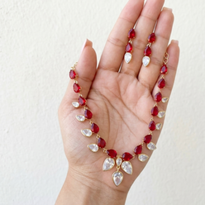 Elegant red gemstone necklace and matching earrings with teardrop-cut red and clear stones set in gold-toned metal, arranged on a luxurious marble background with golden leaf accents.